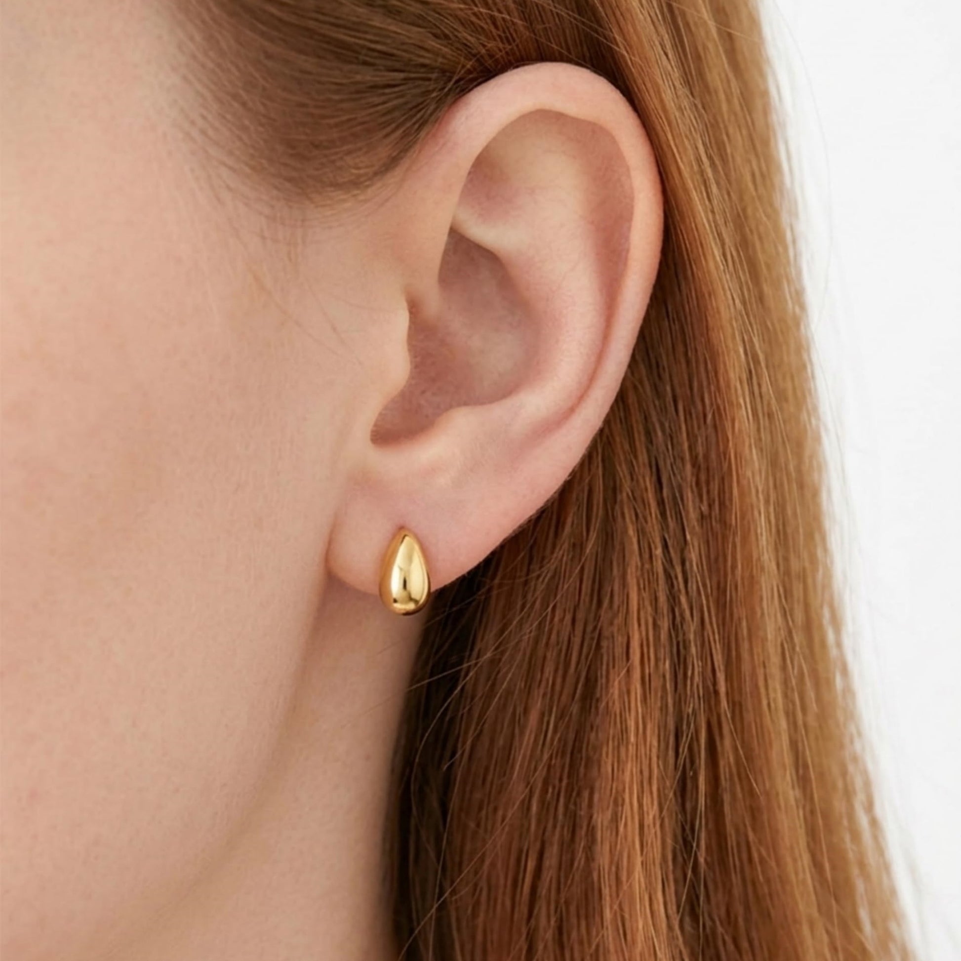 Gold teardrop stud earrings on a model’s ear, smooth oval drop fronts