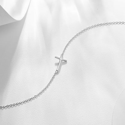 Silver-tone anklet bracelet with sideways cross charm on light fabric.