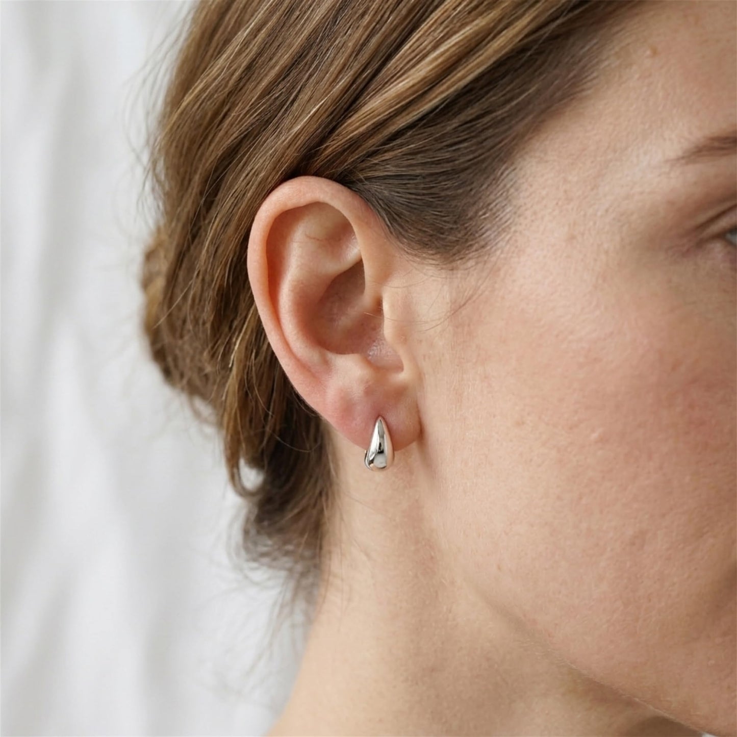 Silver teardrop hoop huggie earring on ear, small curved hoop profile.