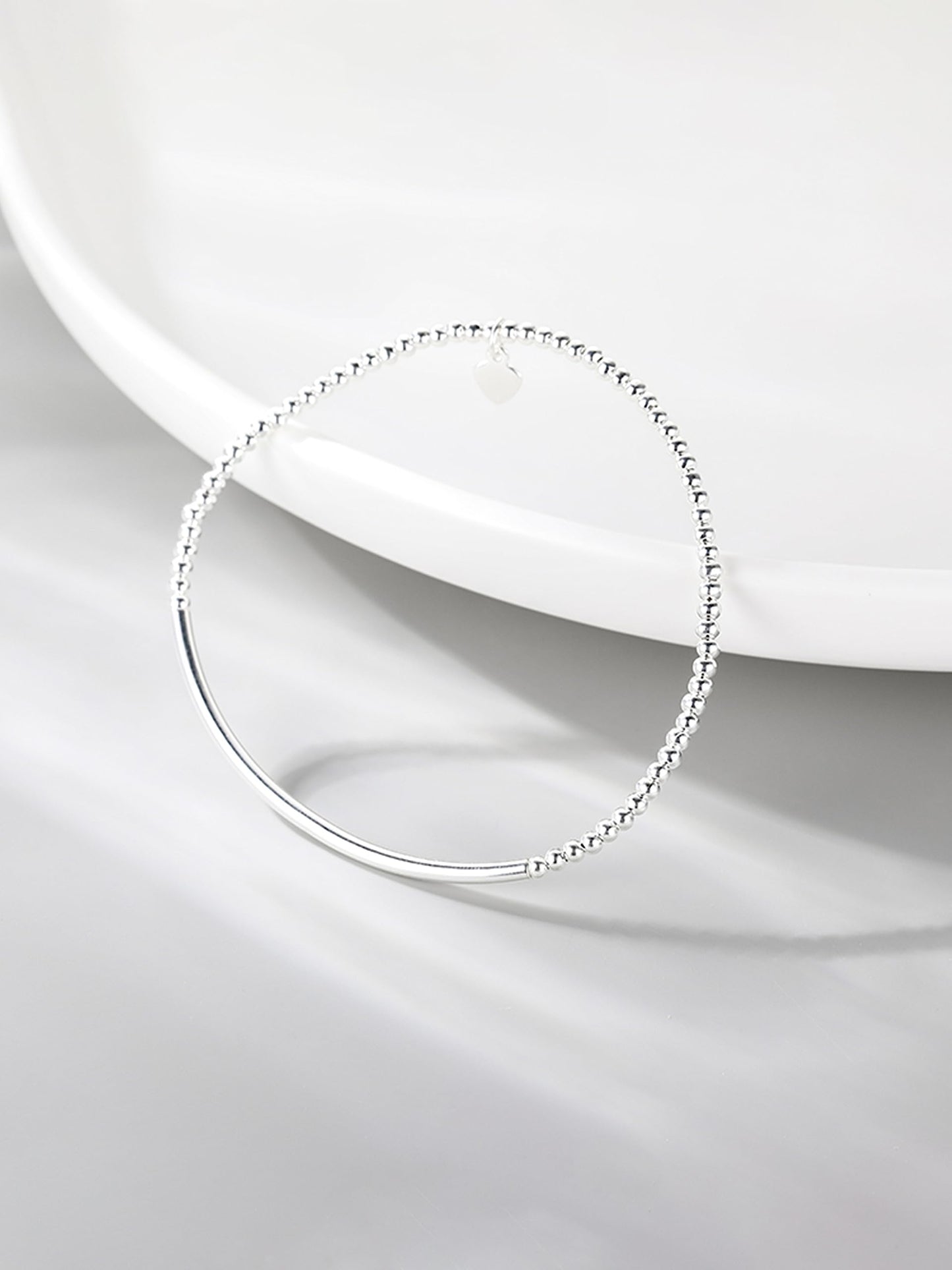 Silver beaded stretch bracelet with heart charm and smooth curved bar on white dish
